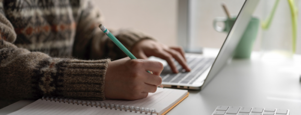 A person sitting in front of a laptop and writing on a notepad with a pencil