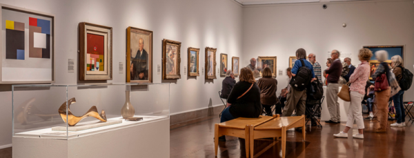 A white gallery filled with modern art. At the end of the room is a group of people listening to a talk. 