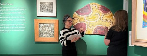 Two women talking in a green art gallery. There is aboriginal style art work hung on the walls.