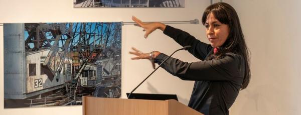 A woman standing at a podium with her arms gesturing to the left hand side. Behind her is a photograph of a historic crane. 