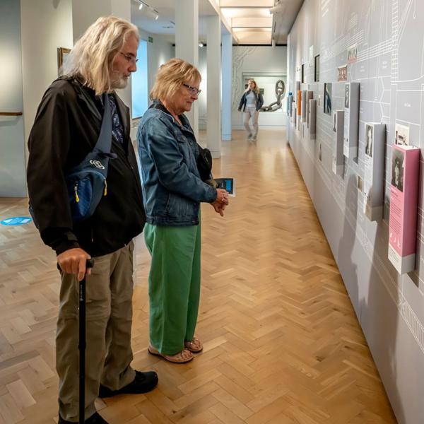 A elderly man and a woman look at a timeline on a wall, the man uses a walking stick. 