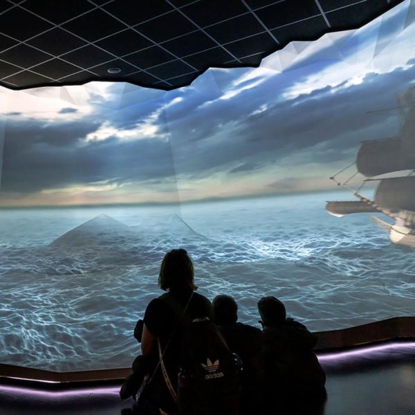 A woman and two children look at a big screen showing a 3D animation of a galleon at sea
