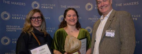 Three people standing in front of a promotional screen which reads 'Heritage Crafts'. On the right is a tall man with grey hair wearing a tweed blazer. On the left is a woman in her 50s with long blonde hair wearing glasses. In the middle is a young woman with curly brown hair wearing a green dress, who is holding an award which looks like a gold sphere made out of textile. 