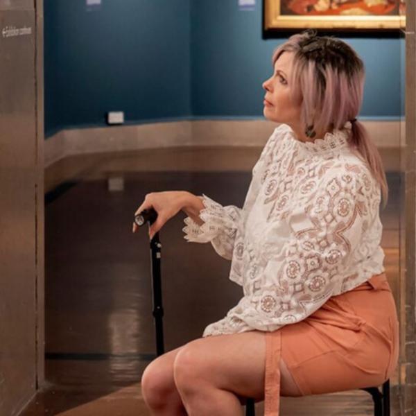 A woman sat on a chair in an art gallery, she has a walking stick by her side. 
