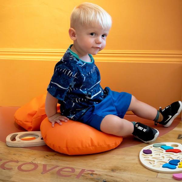 A blonde male toddler sitting on orange cushions on the floor. There are colourful wooden toys around him and the word 'discover' is written on the floor'