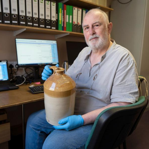 An older man sitting in an office holding a old jug. He is wearing blue latex gloves and looking at the camera. 