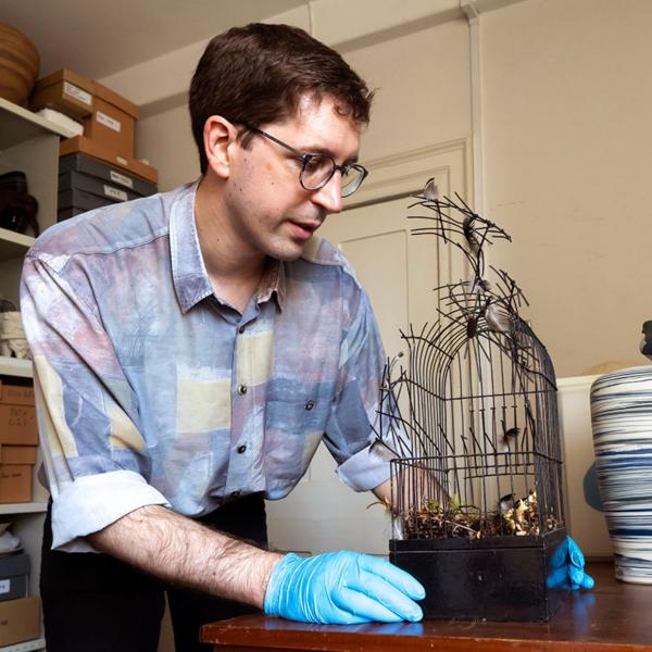 A middle aged man with dark hair and glasses is holding a sculpture. The sculpture looks like a broken bird cage which a bird has just escaped from. 