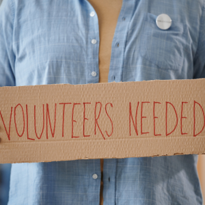 A woman holding a cardboard sign reading 'volunteers needed'