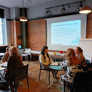 People sitting around round tables in discussion. There is a projector on a wall showing a presentation slide.