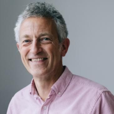 A person (Chris Hollis) with grey hair, wearing a pale shirt, is smiling at the camera