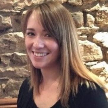 A person (Emma Broome) with long brown hair is smiling at the camera