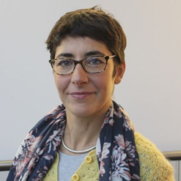 A person with short dark hair and glasses (Elvira Perez Vallejos) is wearing a scarf and smiling at the camera