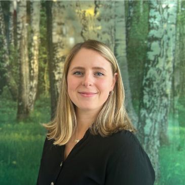 A person (Lucy Hitcham) is smiling at the camera. In the background are trees