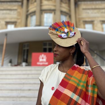 A person (Rianna Patterson) wearing a hat and colourful scarf is standing in front of a building