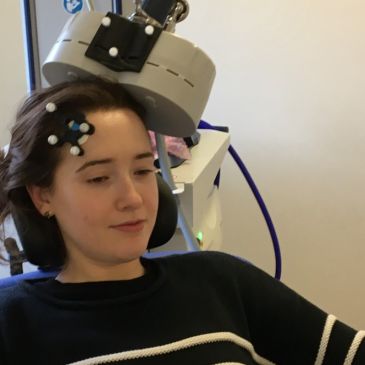 A person is seated whilst receiving transcranial magnetic stimulation. Electrodes are on their head, with a small machine above their head