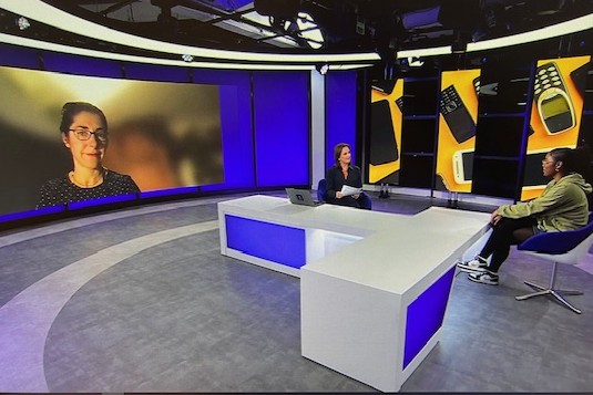 Channel 4 news studio, Elvira speaking on a large screen on the left, news presenter in the centre and a student sat on the right.