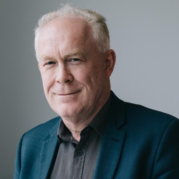 A person is facing the camera and smiling. They're wearing a dark coloured shirt and suit jacket. The background is grey.