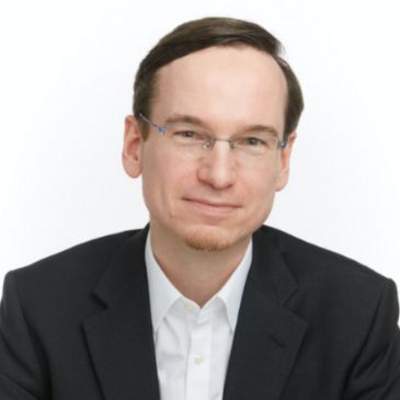 A person with short dark hair and glasses is smiling at the camera. They're wearing a white shirt and black blazer