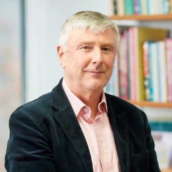 Tom Dening, wearing a pale shirt and dark blazer, smiles at the camera