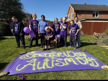 A group of people supported at an Avenues service all wear purple and stand in front of a huge purple sign.