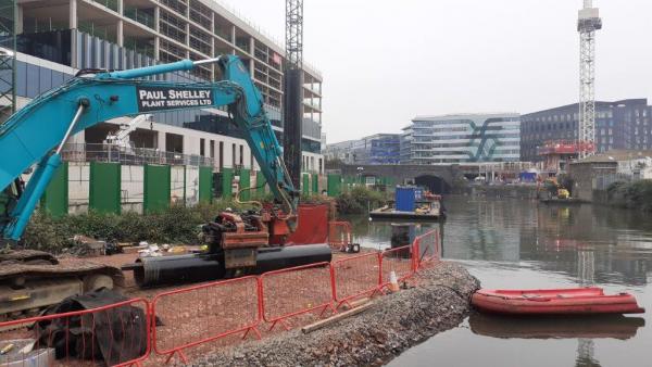 A digger carries out work to install pilings. The digger is on a bed of gravel within the Floating Harbour. 