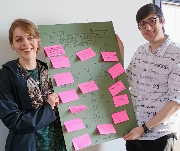 Two people holding up a board which says 'Pledge Tree' with post-it notes stuck on
