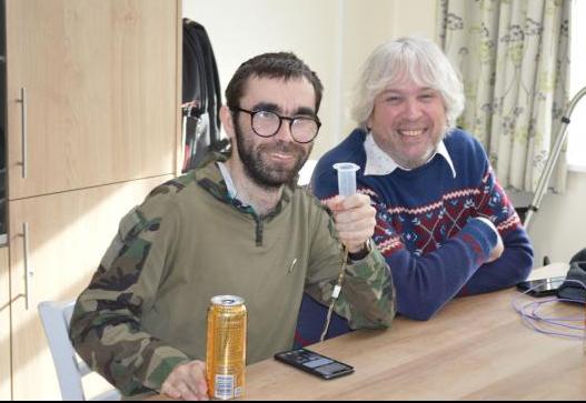 Jacob and his support worker David sit at a table at home smiling at the camera