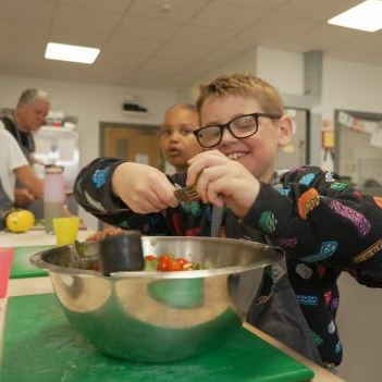 Child wearing glasses mixes ingredients in a bowl during a cooking activity at a table, with other children and adults preparing food together in the background.