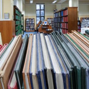 Rows of books on shelves in a library, viewed across a stack in the foreground, with more shelves and reading areas visible in the background.
