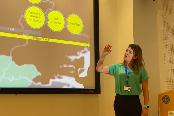 A woman in a green short sleeved shirt and long brown hair presenting at a big screen to the side of her which has a map and some information bubbles on it.