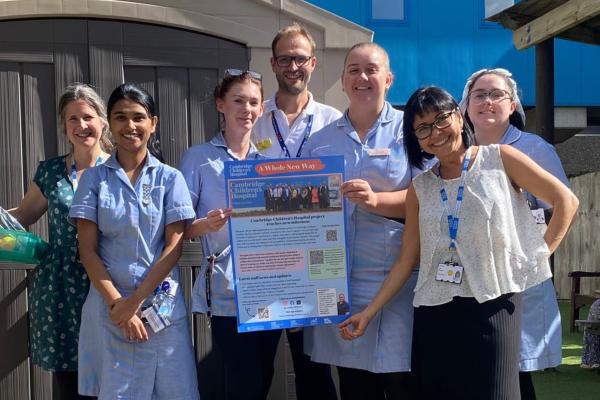 A group of people, mostly in pale blue nurses uniforms, standing in the sunshine holding a colourful cambridge children's hospital poster. They are all smiling
