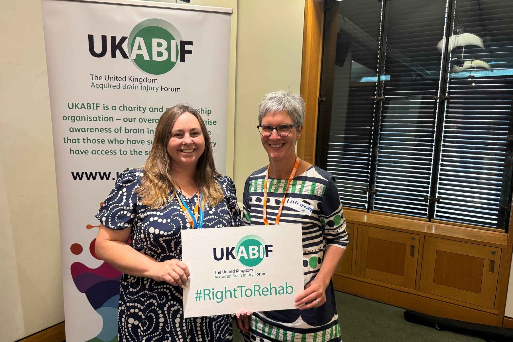 A woman with long brown hair and patterned dress standing next to a woman with short grey hair, glasses, and patterned dress, holding up a placard that says #RightToRehab