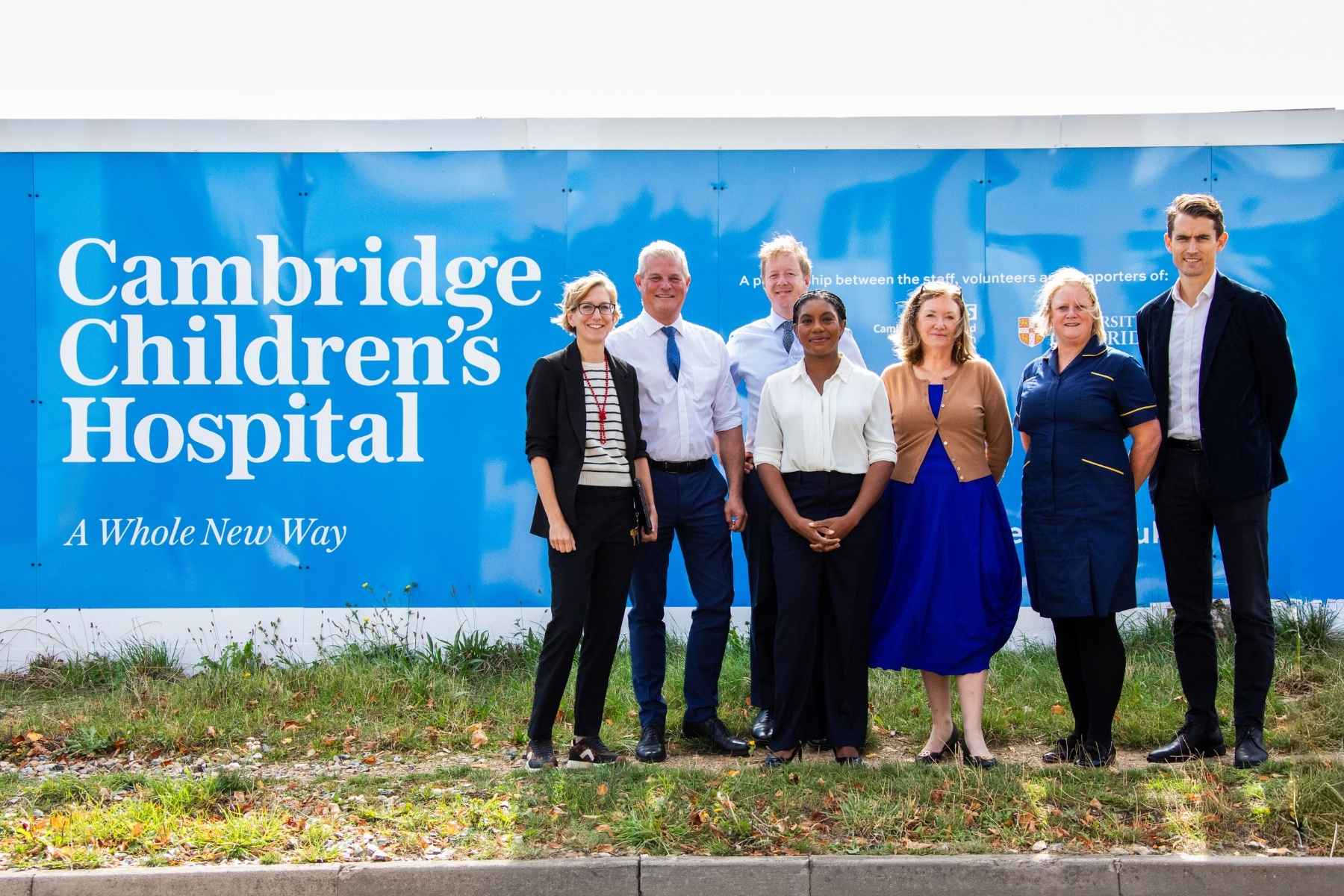 A group of people standing with Kemi Badenoch in front of the Cambridge Children's Hospital hoardings on a sunny day.