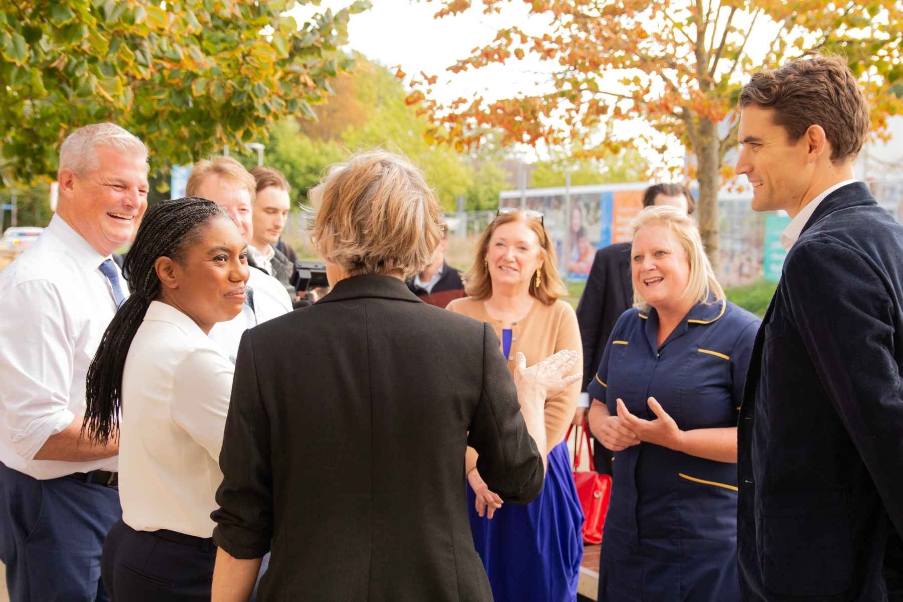 Kemi Badenoch with long braided hair in a pony tail, wearing a smart shirt and standing with a group of people who are talking to her and smiling. One of the people is in nurse's uniform.