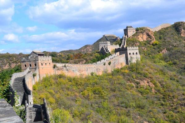 An image of the great wall of china, with its turrets and surrounded by mountainous hillsides and lots of trees and shrubbery