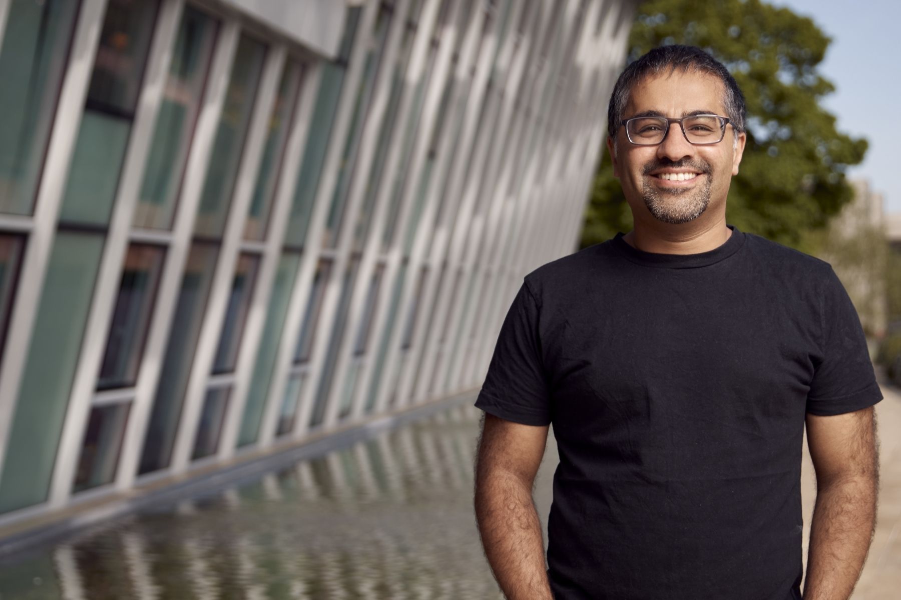 A man with dark grey hair and beard, and dark rimmed glasses, standing in front of a building with a water feature on a sunny day. He is wearing a black t shirt and jeans and is smiling