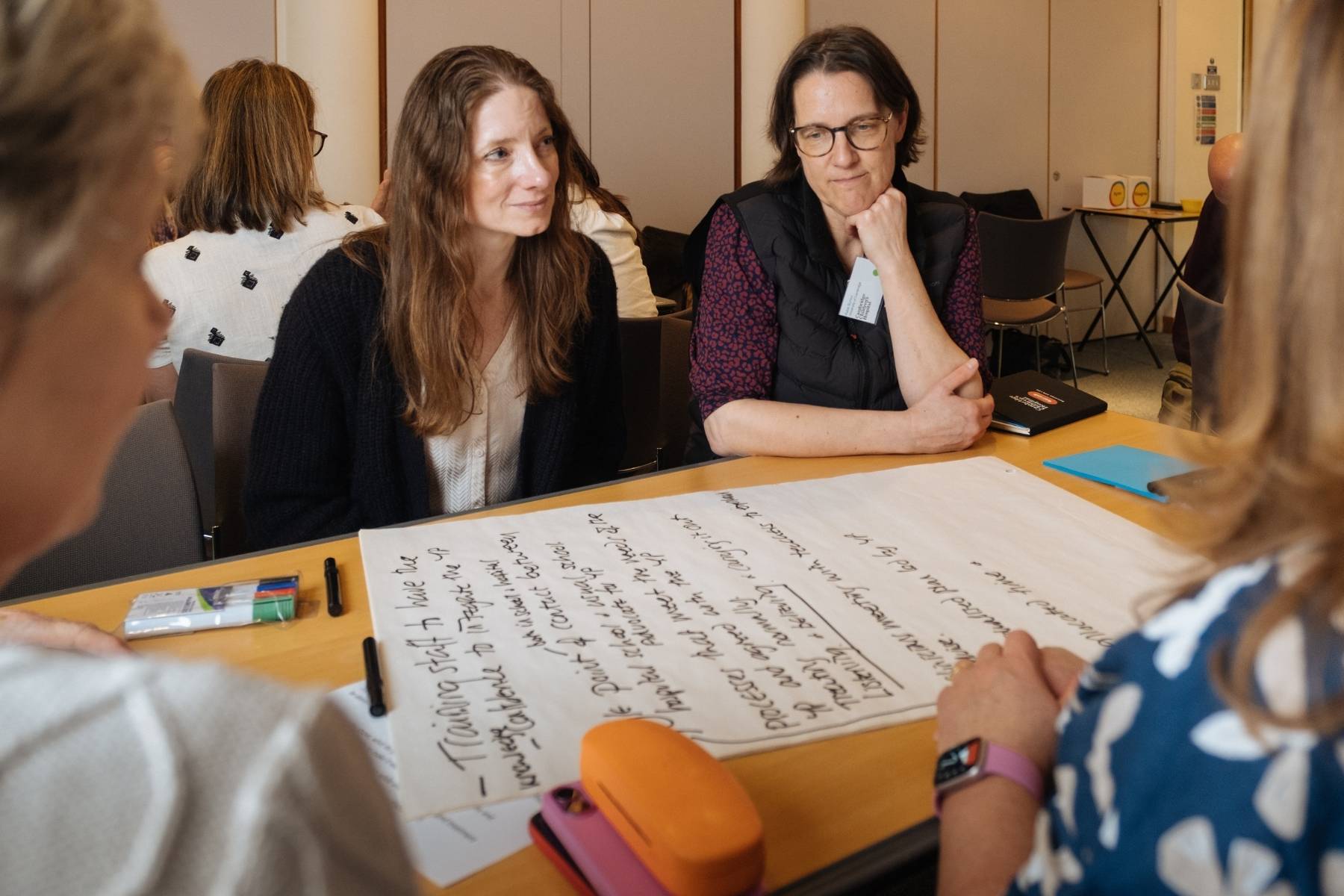 A woman with long brown hair and a woman with shoulder length dark hair and glasses, sitting at a big table with a huge piece of paper in front of them with writing on. This is one of the breakout sessions at the conference.