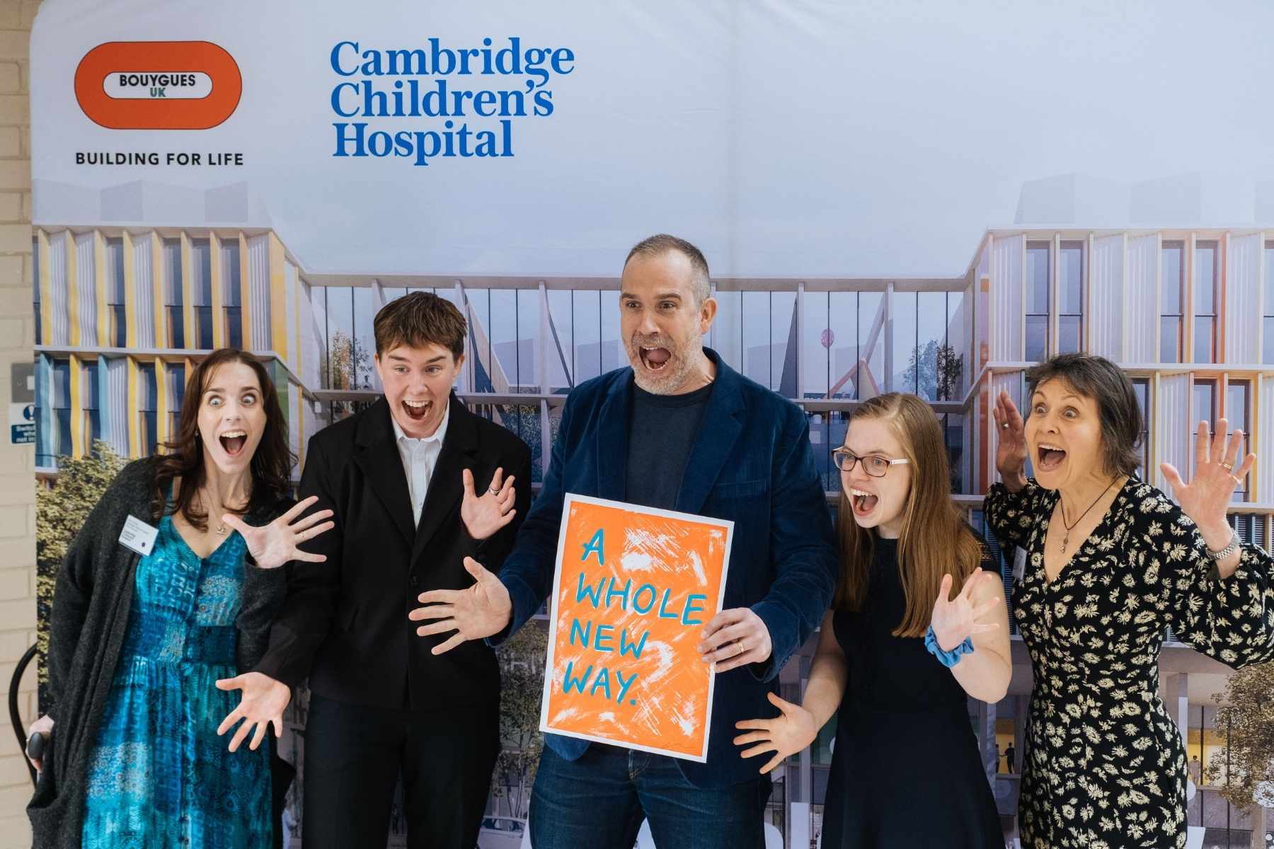 Dr Xand, who has dark grey hair and blue suit, holding an orange poster saying A Whole New Way. Next to him are three women and a young man. They are all laughing and pulling funny faces. 