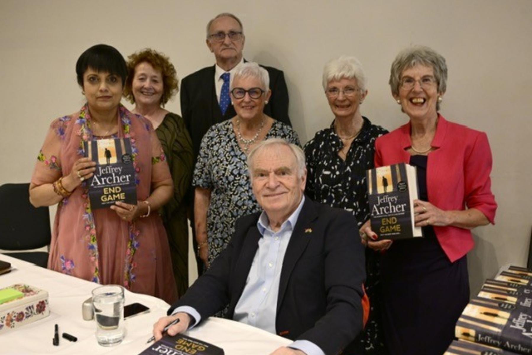 A man with grey hair, pale blue shirt and smart black jacket sitting at a table with piles of books which he is signing. A group of women and a man are standing behind him holding copies of the book. 