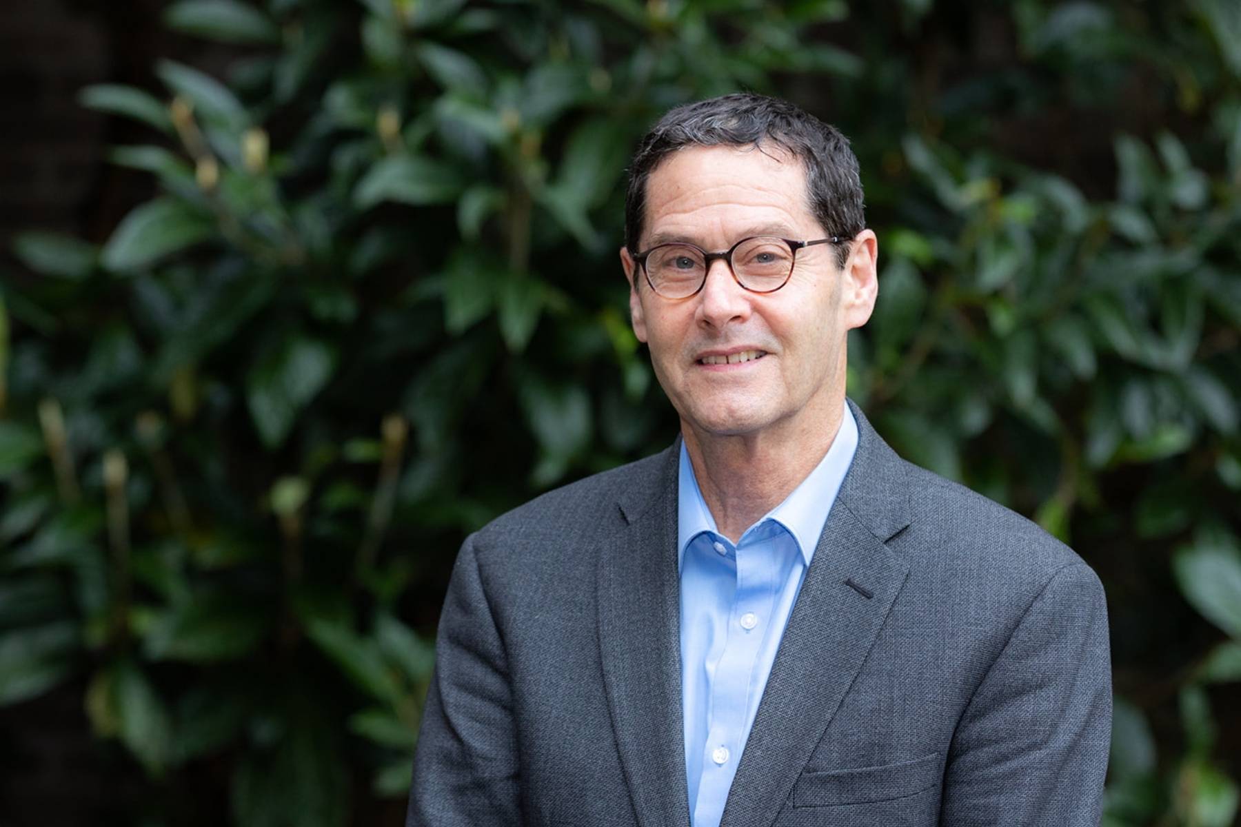 A man with dark short hair, dark rimmed glasses, wearing a pale blue shirt, and dark blue grey suit. He is outside with shrubbery behind him.