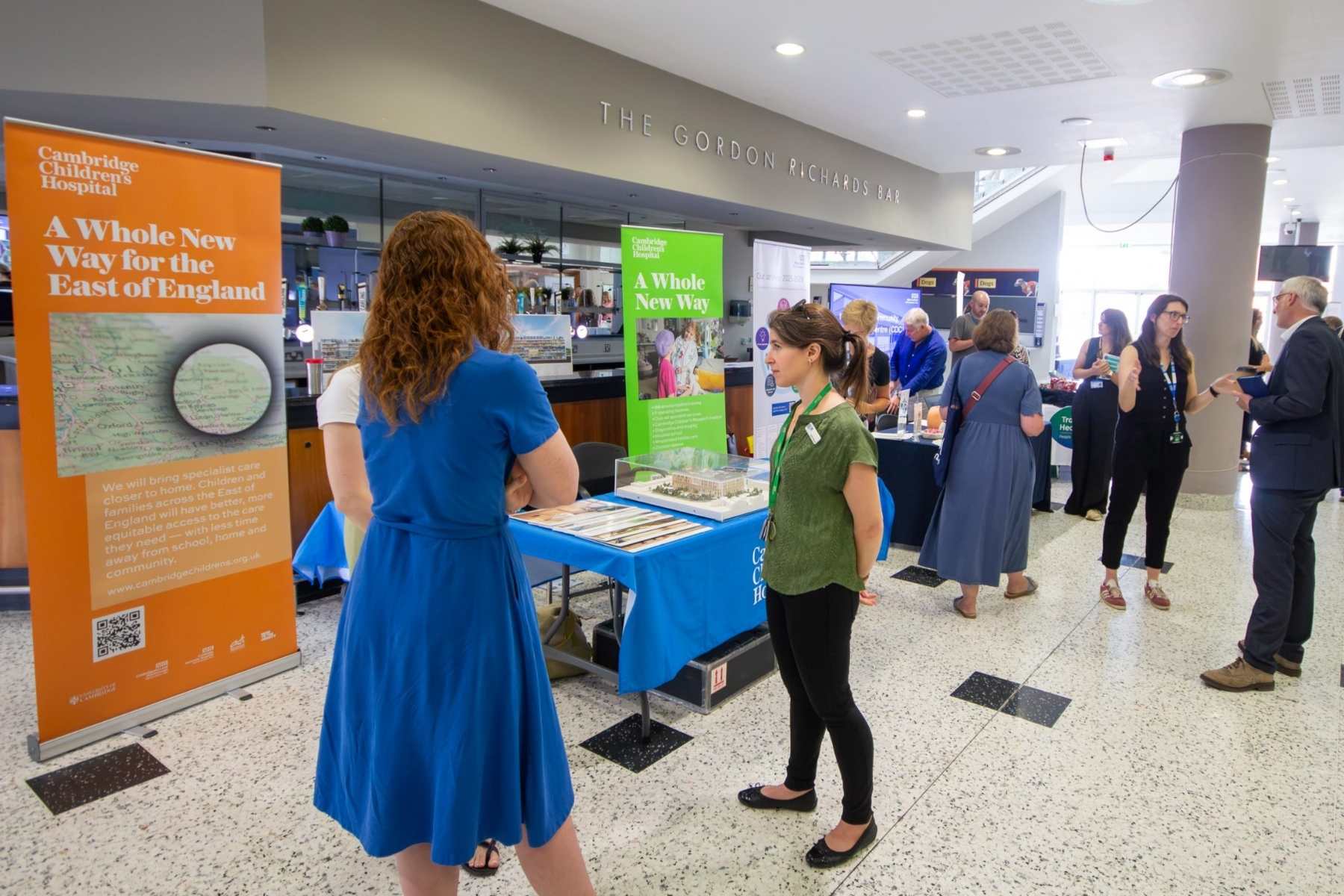 People chatting and looking at stands at a health expo. There are colourful banners for Cambridge Children's Hospital and a table with a blue cloth on it and a model of the children's hospital