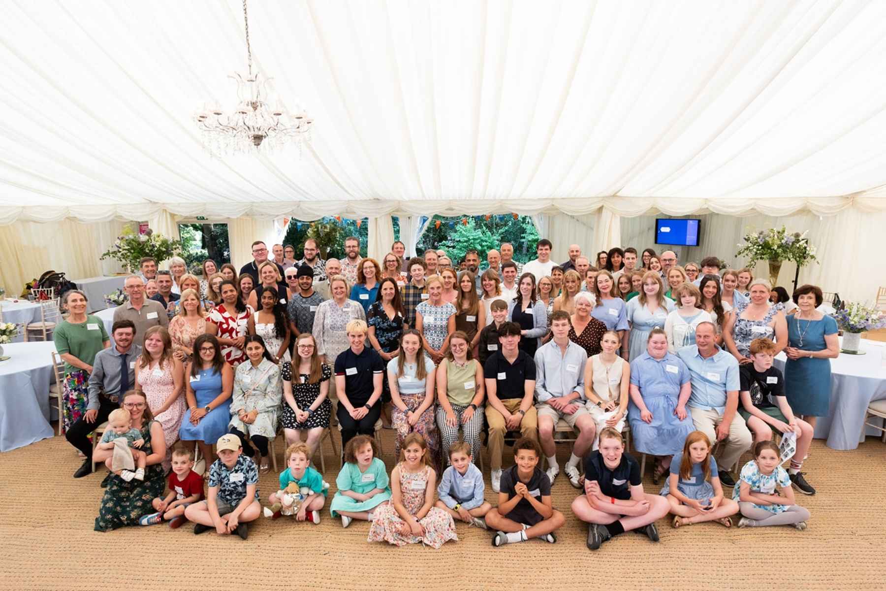 A large group of people, children and adults, standing together in a big fancy marquee. They all look very happy