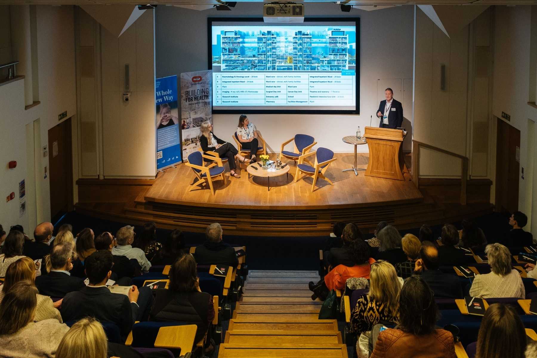 A photo looking down onto the stage from the back of a conference auditorium. The stage is well lit, with people sitting on chairs looking at a man who is standing at a lectern doing a presentation.