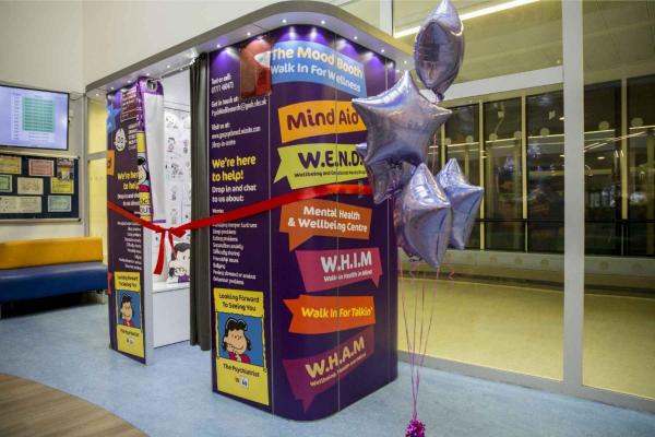 A colourful booth that looks like a photo booth, with an image of Lucy from the Peanuts comic on it. Lucy has black hair and a red top. The booth is surrounded by a red ribbon and balloons. It's in a hospital foyer