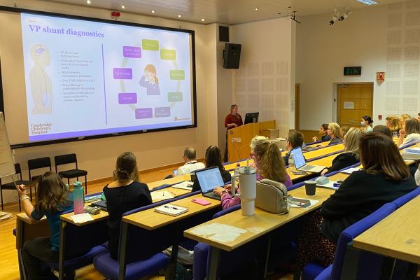 A lecture theatre showing people sitting in the rows of seats, looking down at a colourful slide on a big screen with a woman presenting