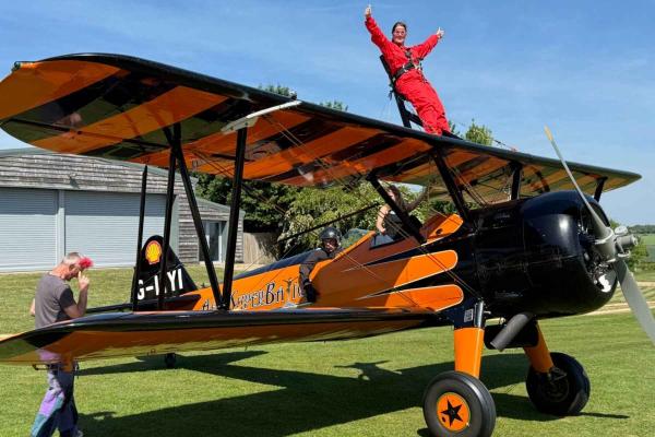 A woman in a red boiler suit standing on the wing of a yellow and black bi plane with her arms outstretched, awaiting take off. She is smiling at the camer and the sky is bright blue