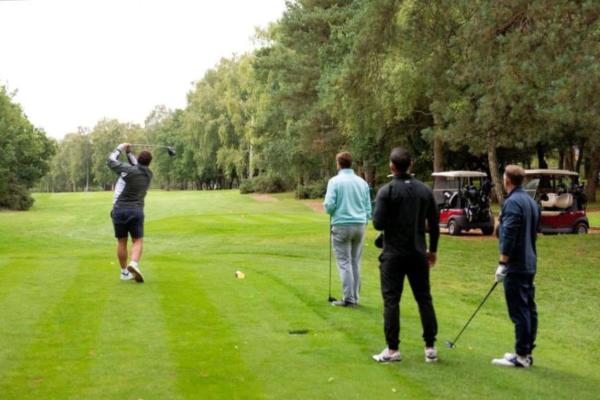 Four men with their backs to the camera looking down a golf green. One man is taking a shot and swinging his club behind him. There are trees and golf buggies on the right hand side.