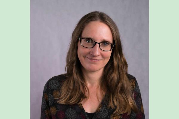 A headshot of a woman with long brown hair, dark rimmed glasses and dark top
