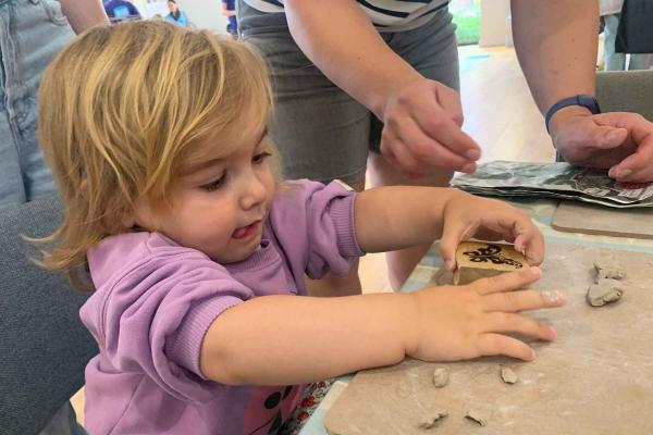 A little girl with fair brown hair and a lilac jumper, playing with a clay activity. she is sticking her tongue out in concentration