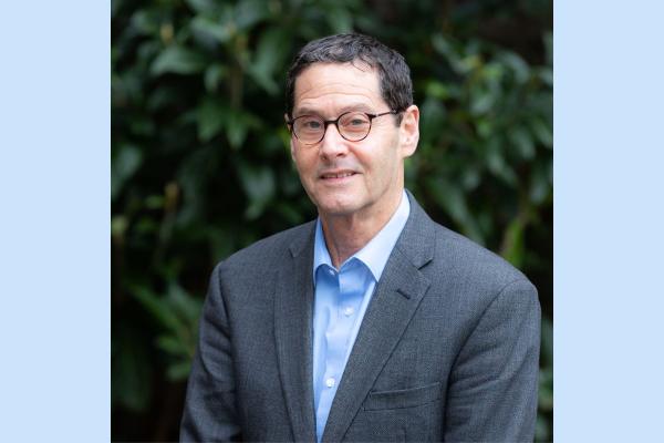 A head and shoulders shot of a man in a dark grey suit, light blue shirt, dark rimmed glasses and dark hair.